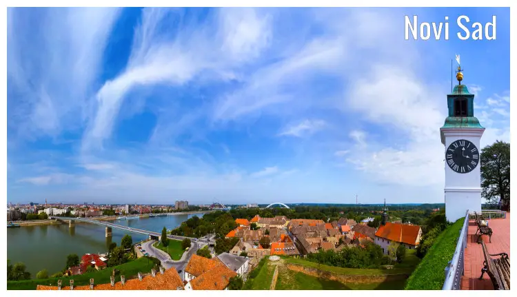 A scenic panorama of Novi Sad, Serbia, with the view of a few buildings, a bridge across the river, and a city clock tower to the right A scenic panorama of Novi Sad, Serbia, with the view of a few buildings, a bridge across the river, and a city clock tower to the right